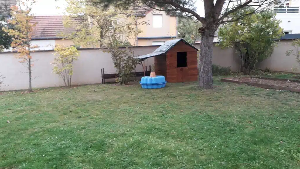 Jardin avec cabane en bois pour enfants, piscine bleue vide, banc et végétation. Mur de séparation en arrière-plan. Ambiance automnale.