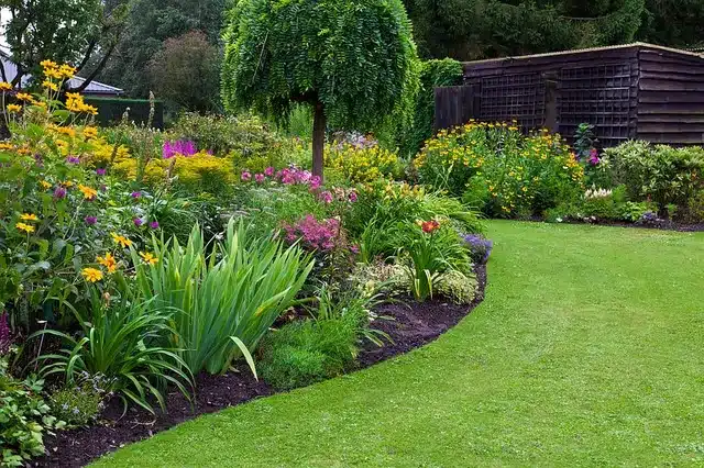 Jardin fleuri avec pelouse verte, arbuste taillé et cabane en bois. Couleurs vives, fleurs jaunes, roses et violettes. Ambiance paisible et verdoyante.
