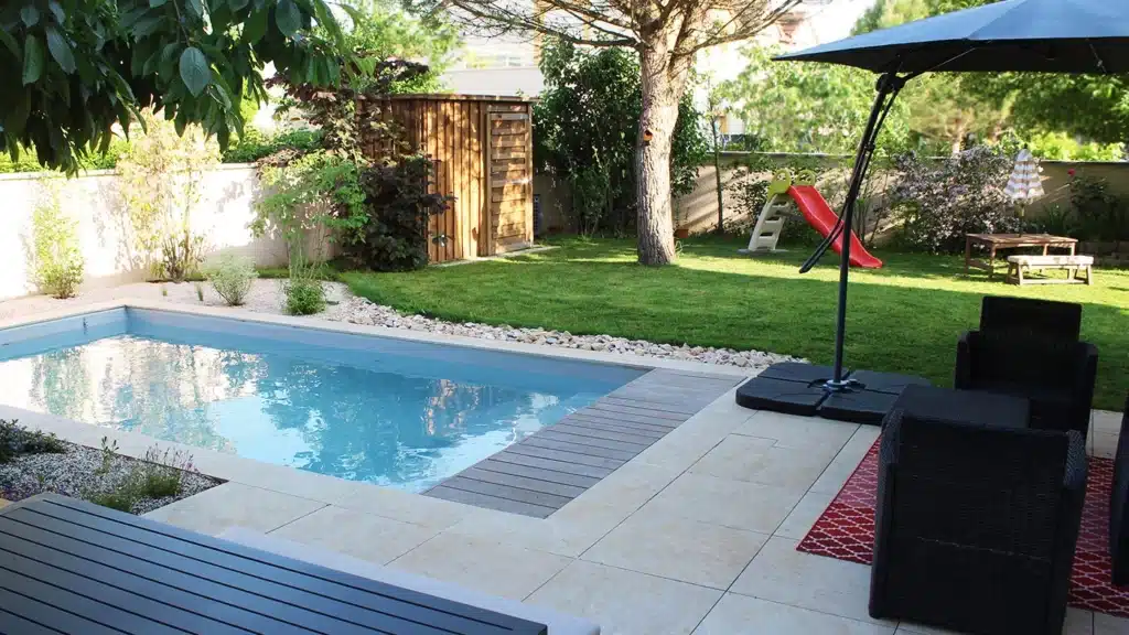 Jardin avec piscine bleue, terrasse dallée et mobilier extérieur noir. Gazon vert, arbre et cabanon en bois au fond. Ambiance paisible.