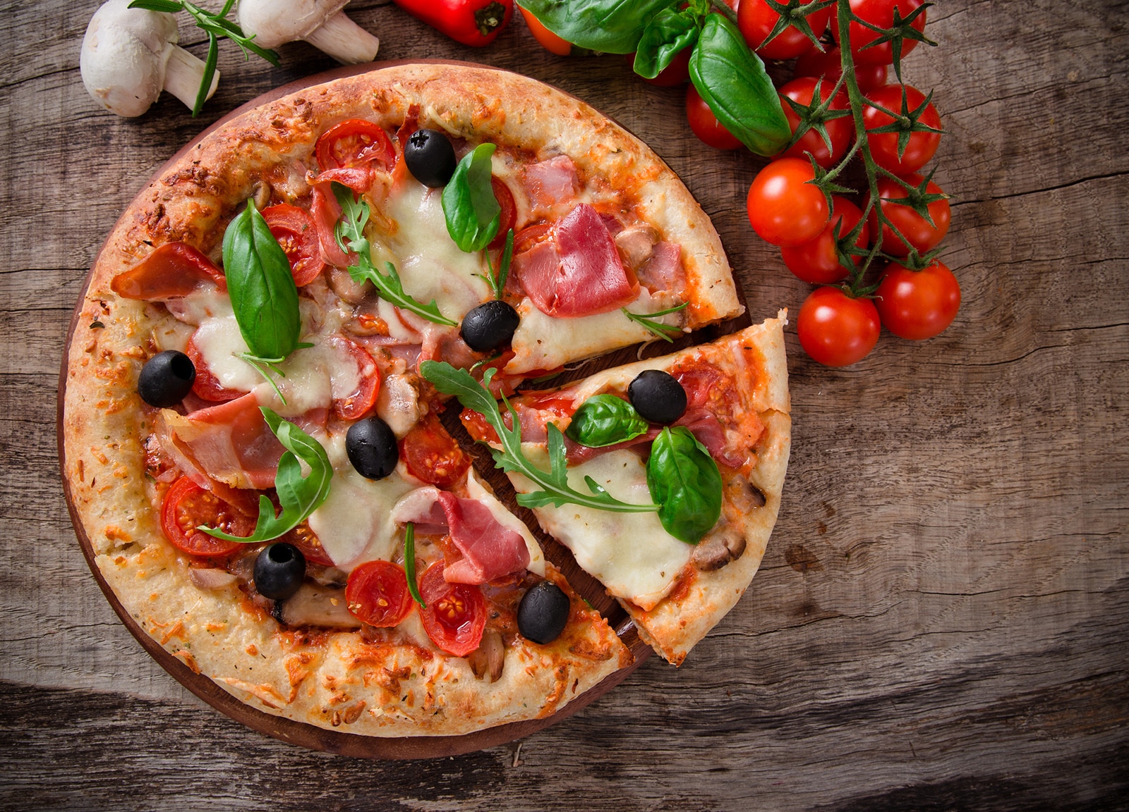 Pizza appétissante avec jambon, tomates, olives et basilic sur une planche en bois rustique. Champignons et tomates cerises à proximité.