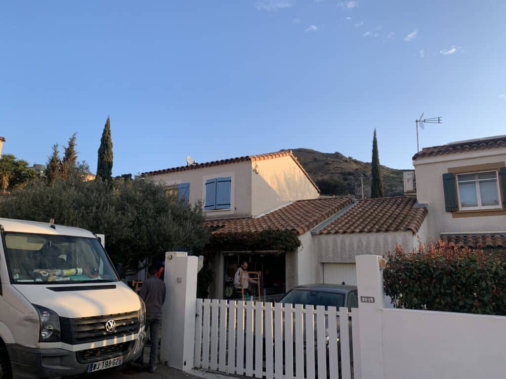 Rue résidentielle méditerranéenne, maisons aux volets bleus et toits en tuiles. Van blanc, deux hommes, ciel bleu. N°12b.