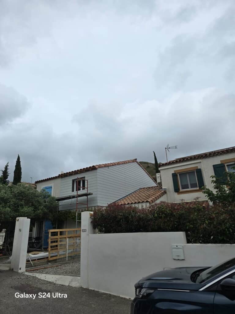 Maison en rénovation avec façade claire et échafaudage sous un ciel gris nuageux. Mur blanc, clôture 32L. Prise Galaxy S24 Ultra.