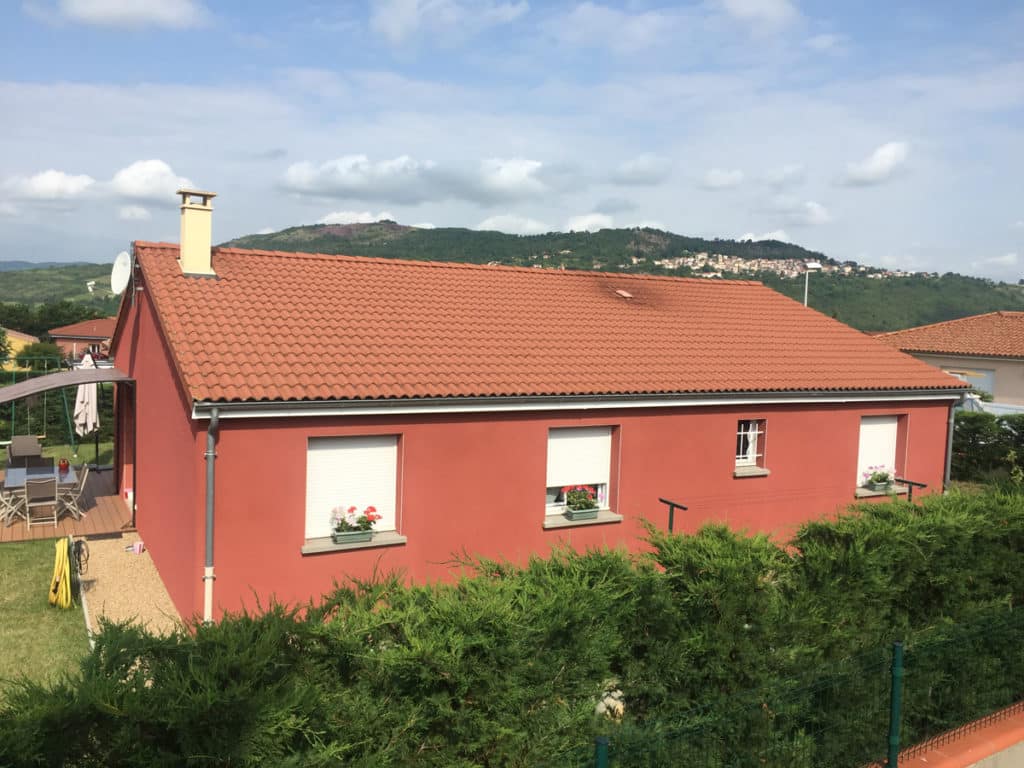 Maison rouge avec toit tuiles, fenêtres volets, jardin, haies, colline avec village et ciel nuageux.