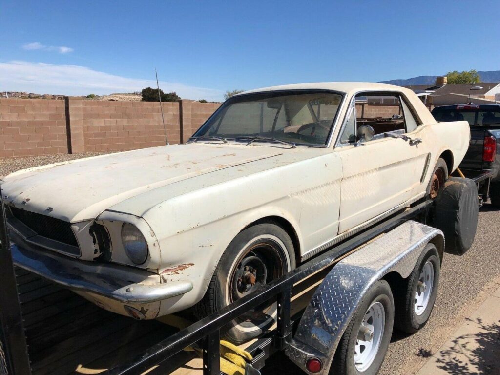 Vieille Ford Mustang coupé blanche rouillée sur remorque, carrosserie usée. Un projet de restauration sous ciel bleu ensoleillé.