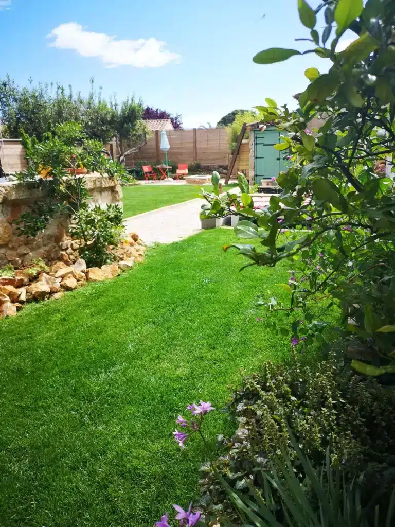 Jardin ensoleillé avec pelouse verte, chemin de gravier et mur en pierre. Cabane verte, mobilier rouge, parasol bleu et fleurs.