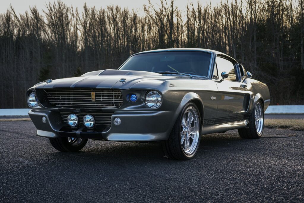 Mustang Shelby GT500 E Eleanor grise avec bandes noires. Vue avant 3/4 sur asphalte, jantes chromées et cobra doré sur calandre.