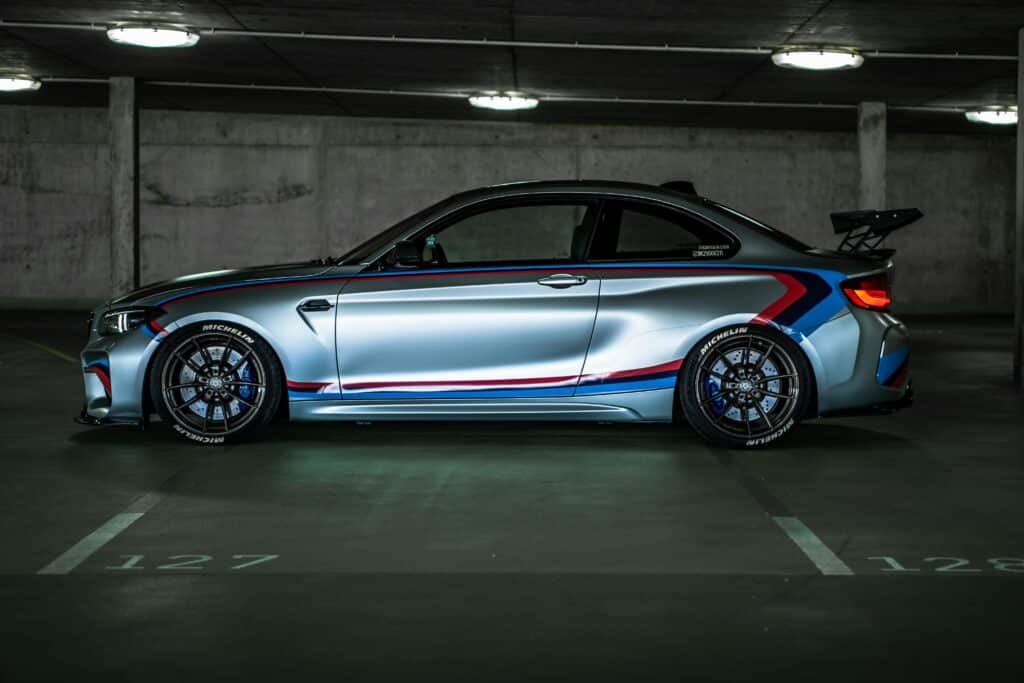 Side view of a BMW M2 sports car with racing stripes, parked in a dimly lit garage.