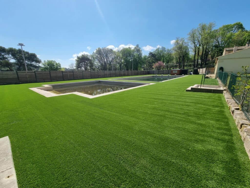 Grand jardin avec gazon synthétique vert vif, deux bassins rectangulaires en béton, clôture en bois et arbres luxuriants sous un ciel bleu clair.