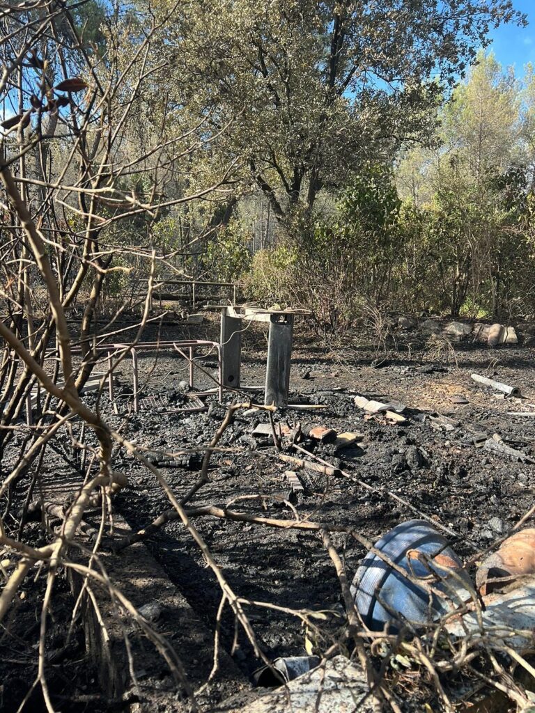 Sol noirci et branches calcinées après un incendie de forêt, avec vestiges de tables et végétation en arrière-plan sous un ciel bleu.